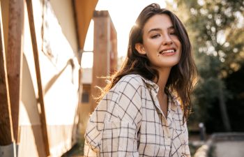 Young woman with long brown hair wearing a plaid shirt smiles outdoors in sunlight, with a blurred background of trees and buildings.