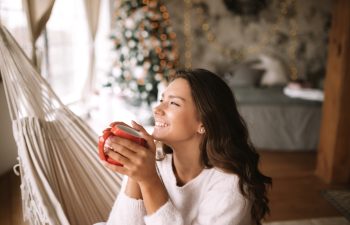 Smiling dark-haired girl dressed in beige sweater and pants holds a red cup sitting in a hammock in a cozy decorated room with a New Year tree